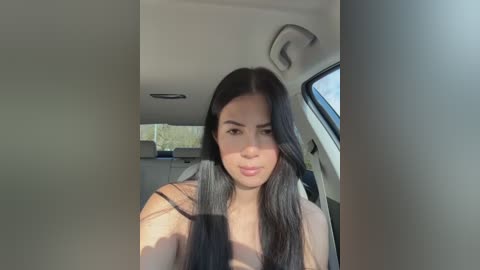 A video of a young woman with long, straight black hair, wearing a beige top, sitting in a car. The background shows the car's interior and a blurred view of greenery outside.