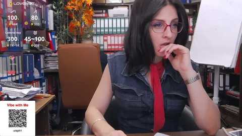 Video of a woman with fair skin, dark hair, glasses, and a red shirt, sitting at a desk in a cluttered office with bookshelves and a floral arrangement.