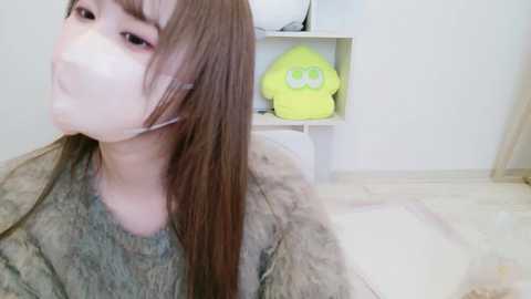 A video of a young woman with long brown hair, wearing a face mask, a green sweater, and a fur coat, seated indoors with a white shelf and yellow plush toy in the background.