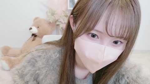 Video of an Asian woman with long brown hair, wearing a pink face mask, light gray sweater, and a beige teddy bear in a soft-focus, pastel-toned room.