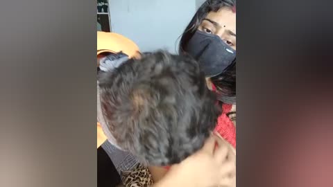Video of a young woman with dark skin, wearing a black face mask, red sweater, and black jacket, hugging a curly-haired child.