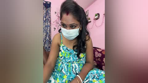 Video of a young woman with dark skin, wearing a blue floral dress and a white face mask, sitting on a bed in a pink room with floral curtains and a wall-mounted lamp.