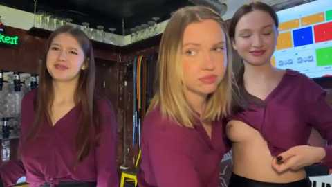 Video of three young women with long, straight hair, dressed in matching purple tops, smiling, standing in a bar with shelves of alcohol in the background.