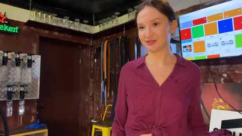 Video of a Caucasian woman with light skin and brown hair tied back, wearing a burgundy blouse, standing in a cluttered, dimly lit room with exposed brick walls, shelves, and a colorful digital display on the right.