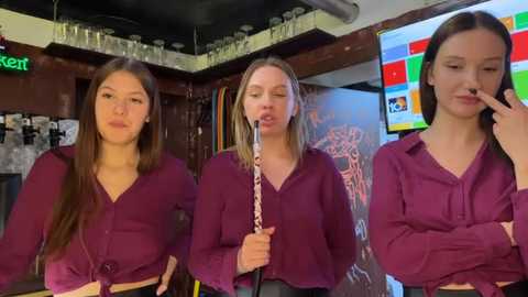 Video of three young women, each with blonde hair, in matching purple shirts, performing on a bar counter. One plays violin, one mimics a violin, and one plays with a finger. Background shows bar shelves and a TV screen.
