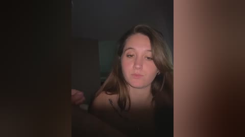 A dimly lit video of a young woman with long, wavy brown hair, wearing a black top, making a pouty face through a door frame.