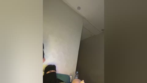 Video of a dimly lit, beige-walled room with a black backpack and a person in a black hoodie, partially visible in the foreground, and a bottle of water on a table.