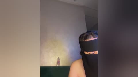 A video of a person with a black blindfold covering their eyes, standing in a dimly lit room with a green wall and a bottle on a shelf.