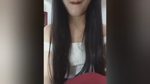 A close-up video of an Asian woman with long, straight black hair, wearing a white, off-shoulder ribbed top, smiling slightly. The background is blurred, showing a red cushion and a white wall.