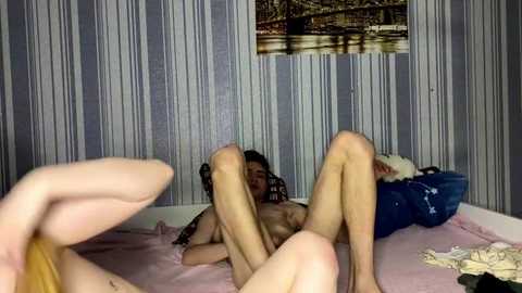 Video of a naked woman with light skin and long black hair, lying on a pink bedspread in a dimly lit room with striped wallpaper. She's holding her legs up, revealing her genitals.