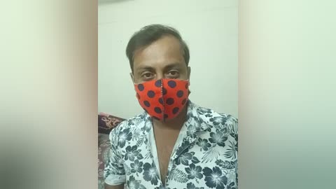 A video of a man wearing a red polka-dotted face mask over a white floral shirt, with a blurred background of a bed and wall.