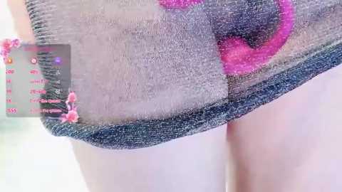 Video of a close-up of a person's lower abdomen, wearing a gray tank top with pink embroidery, featuring a measuring tape with marks and a pink flower pinned to the garment.
