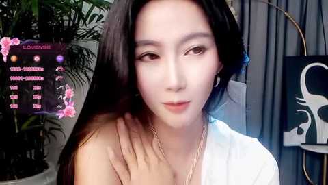 Video of a young Asian woman with fair skin and long black hair, wearing a white top and gold necklace, smiling gently. Background features a green plant and a white and black abstract art piece.