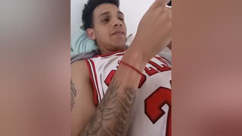 Video of a smiling Latino man with short dark hair, wearing a white and red basketball jersey with number 3, lying on a bed. His tattooed arm is raised.