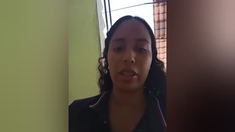 Video of a young Black woman with curly hair, wearing a black shirt, standing in a room with light green walls and a window with brown curtains.