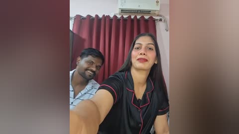 Video of a South Asian woman with medium skin tone, straight black hair, and red lipstick, smiling in a black blouse with red trim. Behind her, a South Asian man with a mustache and glasses, wearing a light blue shirt, smiles. Red curtains and an air conditioner are visible in the background.