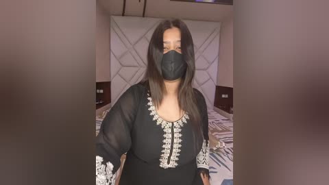 Video of a middle-aged woman with medium skin tone, dark hair, wearing a black face mask, black sheer blouse with white embroidery, and black pants, standing in a bedroom with a white tufted headboard.