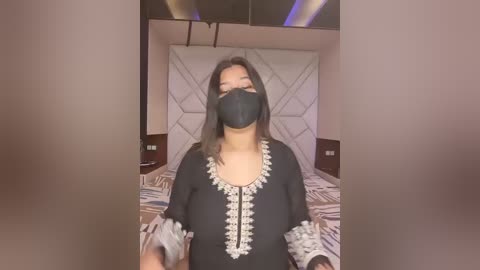 Video of an Asian woman with medium skin tone, wearing a black face mask, black dress with white lace, and black gloves. She stands in a modern bedroom with a geometric-patterned headboard and brown bedding.