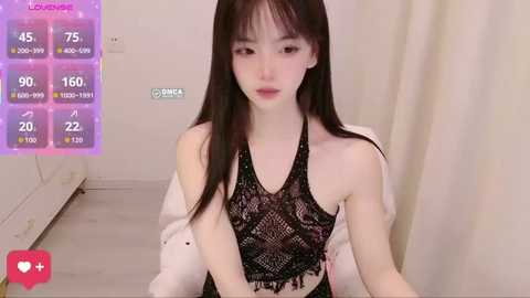 Video of an East Asian woman with fair skin and long black hair, wearing a black lace halter top and white jacket. Background shows a plain room with a purple digital display and white curtains.