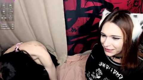 Video of a young woman with straight brown hair and cat ears, wearing a black t-shirt with white text, sitting next to a person tied up on a bed.