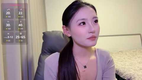 A video of a young Asian woman with long black hair, fair skin, and a neutral expression, wearing a beige cardigan. She sits on a black leather chair in a softly lit room with a beige wall and a fluffy rug.