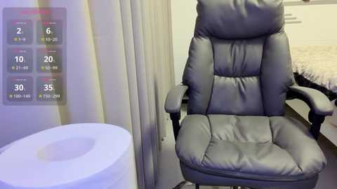 A video of a sleek, gray leather office chair in a modern, minimalist room. To the left, a digital display shows temperature and humidity readings. Background includes a white wall and a bed with white linens.