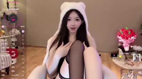 Video of a young East Asian woman with long black hair, wearing a fluffy white hat and black lingerie, sitting on a wooden floor in a cozy, softly lit room.
