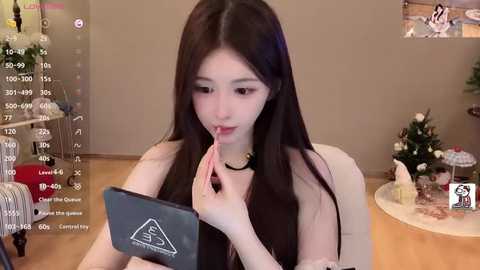 Video of an East Asian woman with long black hair, fair skin, and delicate features, applying lipstick in a cozy, minimalist living room with a Christmas tree in the background.