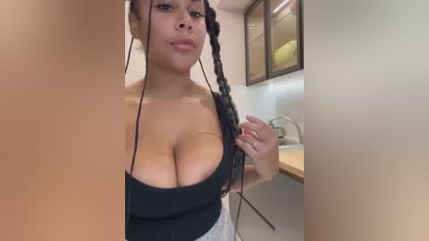 Video of a Black woman with medium skin tone, wearing a black top that reveals ample cleavage, standing in a modern kitchen with beige walls and white cabinets.