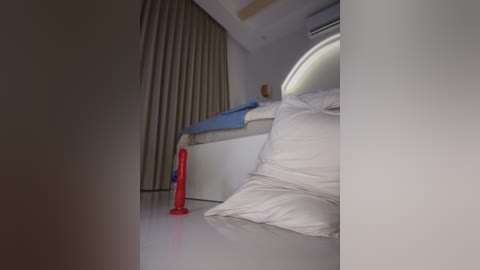 Video of a minimalist bedroom with white walls, beige curtains, a round window, and a white bed with blue and white bedding. A red dildo and a white pillow are on the shiny floor.