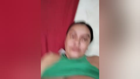 A blurred video featuring a person with dark hair, wearing a green shirt, against a red and white background. The image is out of focus, making details indistinguishable.