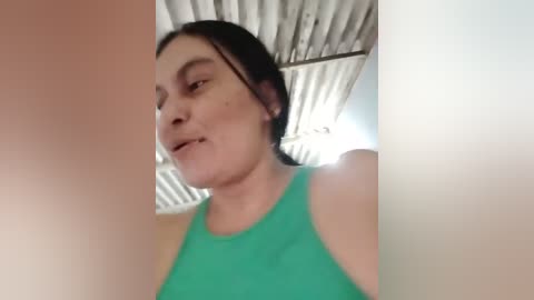 A low-angle, out-of-focus video of a woman with fair skin and dark hair, wearing a green tank top, standing in a dimly lit room with a corrugated metal ceiling.