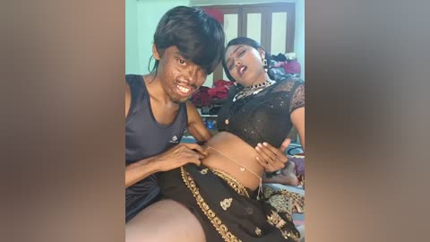 Video of two people: a man with short black hair, dark skin, and a tank top, and a woman with short black hair, fair skin, and a black lace outfit. They are in a bedroom, smiling.