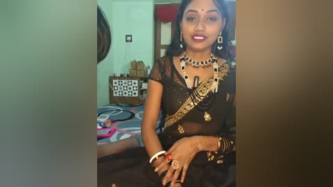 Video of a South Asian woman in a black saree with gold embroidery, adorned with a bold necklace and earrings, sitting in a cluttered, dimly lit room with a white wall and a bed visible in the background.