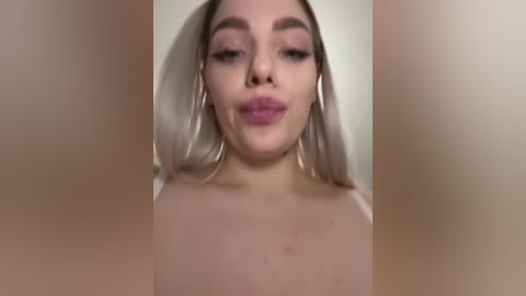 Video of a fair-skinned woman with platinum blonde hair, wearing a white top, puckering her lips in a playful manner. The background is blurred and beige.