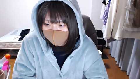 Video of an Asian woman with short black hair, wearing a light blue hooded robe and a beige face mask, sitting at a desk in a cluttered bedroom with a bed, clothes, and a chair visible in the background.