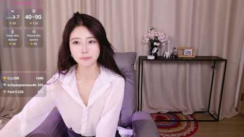 Video of an East Asian woman with long black hair, wearing a white blouse, seated in a gray chair, surrounded by a floral arrangement and digital display.