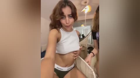 Video of a young, light-skinned woman with curly brown hair, wearing a white crop top and black Calvin Klein underwear, sitting on a white laundry basket in a cluttered, dimly lit room.