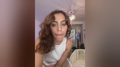 Video of a young woman with wavy brown hair, light skin, and a white tank top, posing in a modern, beige-toned living room with a ceiling fan, a potted plant, and a white basket.