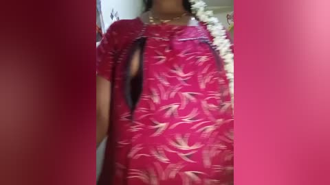 Video of a woman in a pink floral dress with a large, deep V-neckline, partially revealing her black bra. She wears a pearl necklace and a floral garland around her neck. The background is a red wall.