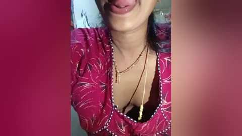 Video of a smiling woman with medium brown skin, wearing a pink blouse with white floral patterns, a gold necklace, and a beaded choker. The background is blurred and magenta-colored.