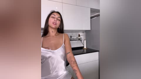 Video of a Latina woman with long dark hair, wearing a white camisole, leaning against a modern kitchen with white cabinets and a black countertop. She has a relaxed, sensual expression.