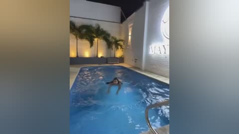 Video of a woman swimming in a lit indoor pool with white walls, palm trees, and \"MAX\" sign in the background.
