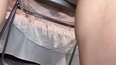 Video of a naked person with fair skin sitting on a chair with a glass top, white sheets visible beneath.