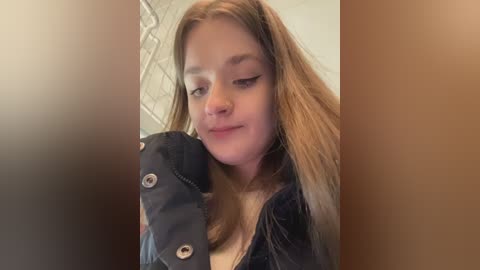 Video of a young woman with light skin and long, straight, blonde hair, wearing a dark jacket, with a slight smile. Background features a blurred, beige wall and a faint geometric pattern.