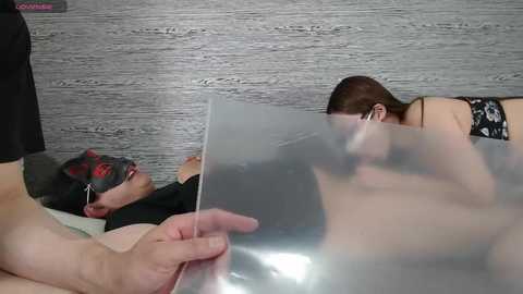 Video of a man wearing a black mask and a black shirt, holding a transparent barrier. Behind him, a woman in black lingerie and glasses lies on a bed with a gray, textured wall background.