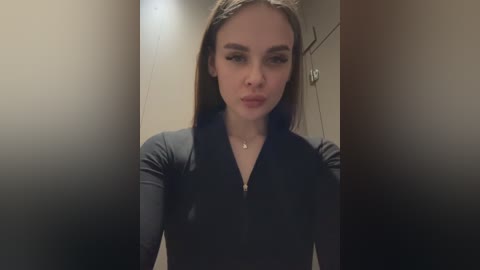 Video of a fair-skinned woman with straight, medium-length brown hair, wearing a black, long-sleeved top. She stands in a dimly lit, beige hallway with blurred, darkened background.