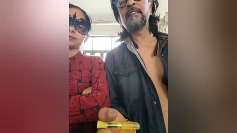 Media: Video of a man with long hair and a beard, wearing a black mask and a red-patterned shirt, holding a yellow handgun, standing next to a woman in a black mask and a red-patterned top.