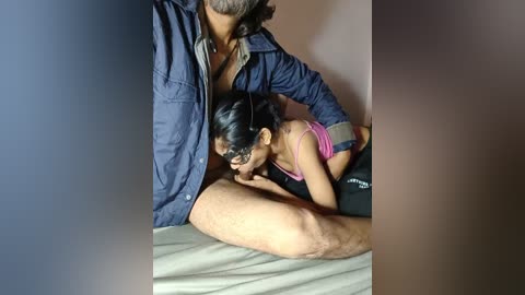 Video of a man with gray hair in a blue shirt, sitting on a bed, with a woman with black hair and glasses performing oral sex on him.