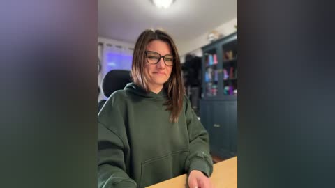 Video of a woman with glasses and shoulder-length brown hair, wearing a dark green hoodie, sitting at a desk in a dimly lit room with a bookshelf in the background.